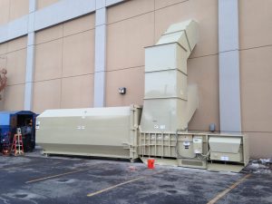 A large beige stationary compactor positioned against a building, featuring a chute extending upwards, with an orange bucket and blue tarp situated nearby.