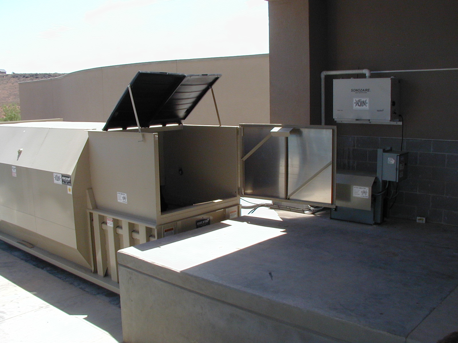Large rooftop self-contained compactor with open maintenance panels, installed on a concrete platform beside a building wall.