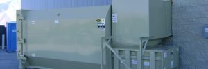 Large industrial waste compactor next to a wall, with a blue dumpster in the background on a concrete surface.