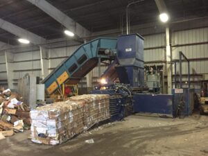 Industrial recycling machine compresses cardboard into bales inside a warehouse with surrounding loose materials and conveyor belt.