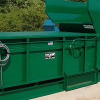 An outdoor installation features a sizable green Stationary Compactor positioned on a concrete surface, set against a backdrop of trees.