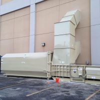 A large beige stationary compactor positioned against a building, featuring a chute extending upwards, with an orange bucket and blue tarp situated nearby.