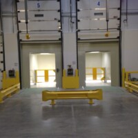 Interior view of a loading dock area equipped with Stationary Compactors, featuring three numbered bay doors, safety barriers, and a concrete floor.
