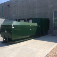 A green Stationary Compactor is positioned outside a building next to a UPS truck.
