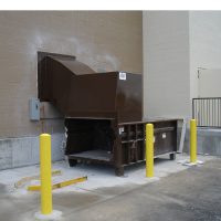 A sizable Stationary Compactor with a brown finish is positioned against a beige wall, encircled by yellow bollards on a concrete surface.