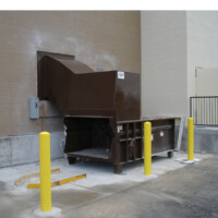 A sizable Stationary Compactor with a brown finish is positioned against a beige wall, encircled by yellow bollards on a concrete surface.
