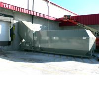 A Stationary Compactor is situated outside a building near a loading dock, featuring a red roof and with a crane visible in the background.