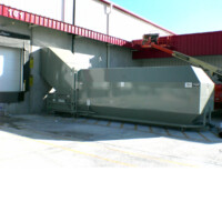 A Stationary Compactor is situated outside a building near a loading dock, featuring a red roof and with a crane visible in the background.