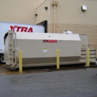 Large white industrial waste compactor in an alley, with yellow safety bollards in front and a truck in the background.