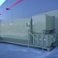 Large industrial waste compactor next to a wall, with a blue dumpster in the background on a concrete surface.