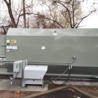 Large gray industrial compactor outdoors, surrounded by trees and power lines, with a small control panel on the left.