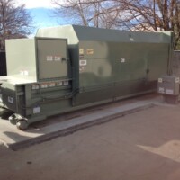 Large industrial trash compactor on a concrete pad, surrounded by a metal fence and trees in the background.
