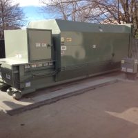Large industrial trash compactor on a concrete pad, surrounded by a metal fence and trees in the background.