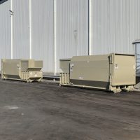 Two large beige dumpsters sit outside near a white industrial building on a paved surface.