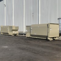 Two large beige dumpsters sit outside near a white industrial building on a paved surface.