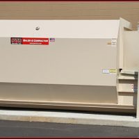 Large industrial waste compactor installed outside a building, next to a utility hookup, on a paved ground surface.