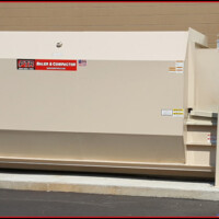 Large industrial waste compactor installed outside a building, next to a utility hookup, on a paved ground surface.