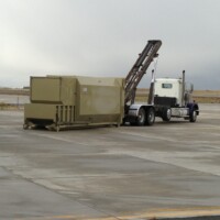 A semi-truck is positioned on a concrete surface, equipped with a long-arm mechanism to lift a large green container.
