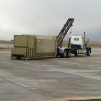 A semi-truck is positioned on a concrete surface, equipped with a long-arm mechanism to lift a large green container.