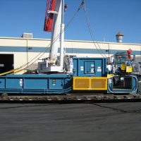 A large industrial machine loaded on a flatbed truck near a FedEx building under a clear blue sky.