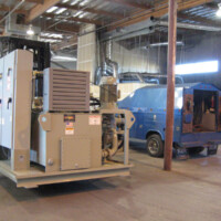 A large industrial machine near a blue van inside a warehouse with exposed beams and ductwork.