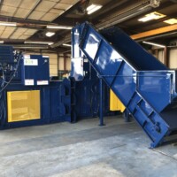 A large industrial blue compactor machine with an attached conveyor belt is in a warehouse setting.