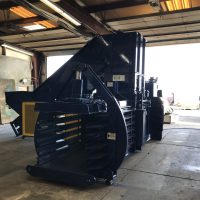 A large industrial baler machine sits inside a warehouse with an open garage door.