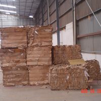Stacks of compressed cardboard bales are arranged inside a large warehouse with a high ceiling.