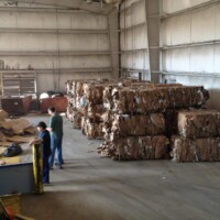Warehouse with large bales of compressed cardboard, two people sorting materials, and machinery visible in the background.