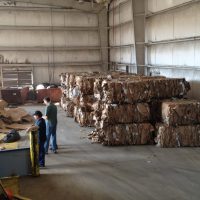Warehouse with large bales of compressed cardboard, two people sorting materials, and machinery visible in the background.