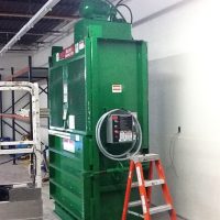 A green industrial compactor in a warehouse setting with a red step ladder positioned beside it.