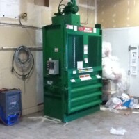 Green industrial compactor in a utility room with bags of waste nearby and various electrical equipment on the walls.