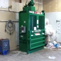 Green industrial compactor in a utility room with bags of waste nearby and various electrical equipment on the walls.