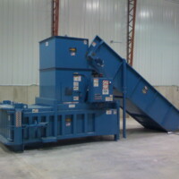 A large blue industrial baler machine with a conveyor belt is situated in a spacious warehouse with concrete walls.