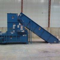 A large blue industrial cardboard baler with a conveyor belt in a warehouse setting.
