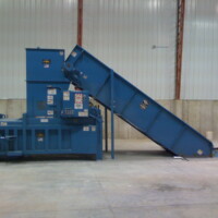 A large blue industrial cardboard baler with a conveyor belt in a warehouse setting.