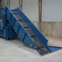 A blue industrial conveyor belt in a warehouse, with a ramp leading into a machine for processing materials.