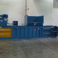 A large blue industrial compactor machine with a hopper is situated inside a warehouse with a plain white wall.