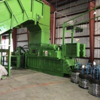 A large green industrial baler in a warehouse, surrounded by coiled wire and conveyor belts.
