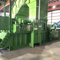 A large green industrial baler machine in a warehouse setting, used for compacting materials.