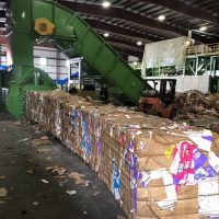 Bales of compacted cardboard stacked in a recycling facility with a conveyor belt and forklift nearby.