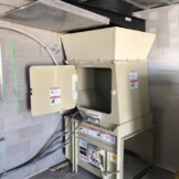 Industrial cardboard baler in a warehouse setting with the loading door open, surrounded by concrete walls and piping.
