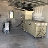 Industrial waste compactor in a concrete room, connected to electrical control panels and secured to the wall.