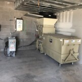 Industrial waste compactor in a concrete room, connected to electrical control panels and secured to the wall.