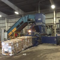 Industrial recycling machine compresses cardboard into bales inside a warehouse with surrounding loose materials and conveyor belt.