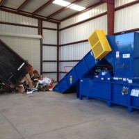 A waste disposal truck unloads recycling materials into a blue industrial compactor inside a warehouse.