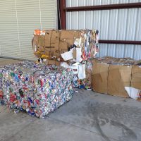 Three compacted bales of recyclable material, including cardboard and mixed recyclables, are stacked in a metal building.