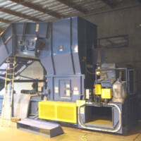 Industrial baler machine in a warehouse setting with a surrounding ladder and safety equipment.