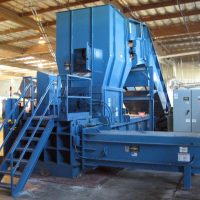 A large industrial baler machine in a warehouse setting, featuring steps, railings, and control panels.