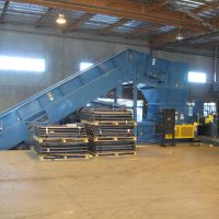 Large industrial blue metal conveyor system in a warehouse, with stacked materials in the foreground and a control panel nearby.